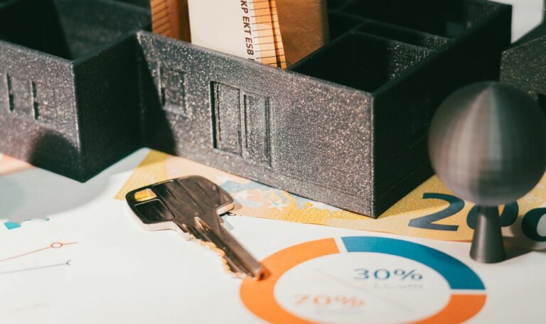 Image macro d'une scène sur une feuille de statistiques et de graphiques financiers. Au centre, une maquette de maison sombre et une clé dorée reposent sur des billets de banque en euros. Deux petites formes d'arbres métalliques sont visibles. Ce visuel évoque l'analyse financière et la Rétrospective marketing tendances 2025, notamment l'investissement et les résultats économiques.