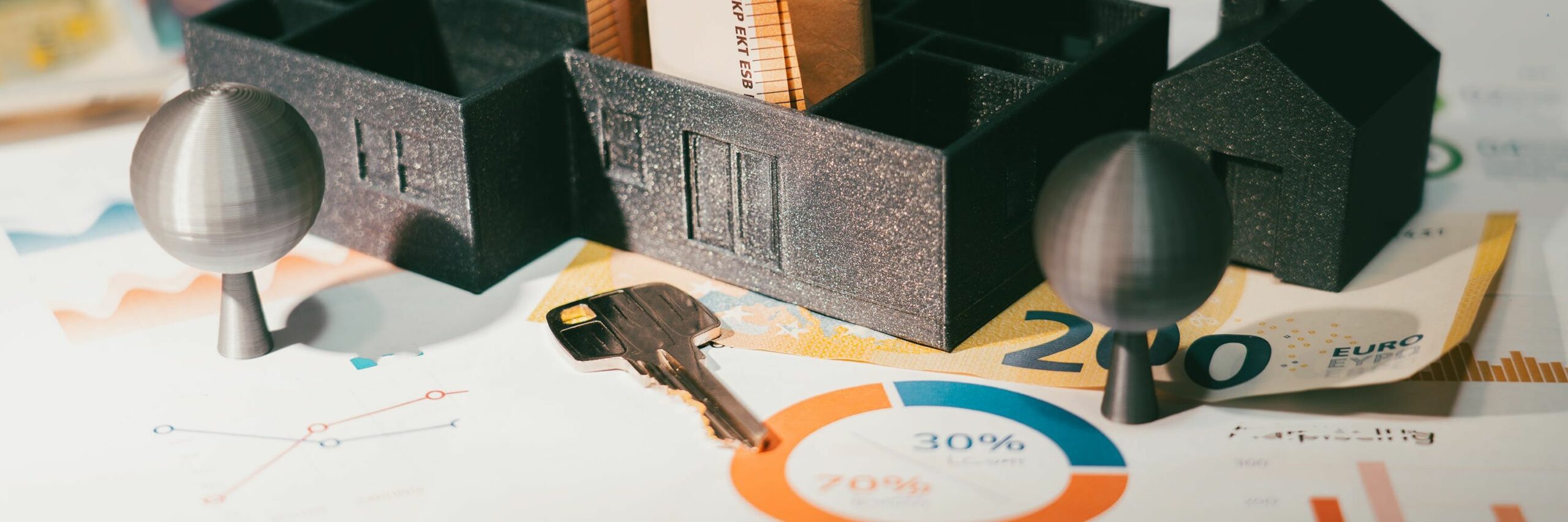 Image macro d'une scène sur une feuille de statistiques et de graphiques financiers. Au centre, une maquette de maison sombre et une clé dorée reposent sur des billets de banque en euros. Deux petites formes d'arbres métalliques sont visibles. Ce visuel évoque l'analyse financière et la Rétrospective marketing tendances 2025, notamment l'investissement et les résultats économiques.