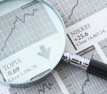 Gros plan sur une loupe grossissant un journal financier ouvert. Sous la loupe, un graphique boursier monte en flèche, avec un texte affichant l'indice 'TOPIX -0.69 Close 1,196.78' et une flèche pointant vers le bas, indiquant une tendance à la baisse locale. À côté, l'indice 'NIKKEI +25.6 Close 14,467.14' apparaît avec une flèche pointant vers le haut, symbolisant l'analyse des tendances économiques et des marchés financiers.