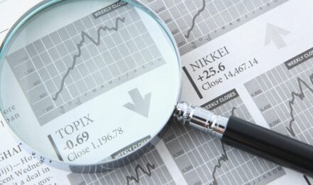 Gros plan sur une loupe grossissant un journal financier ouvert. Sous la loupe, un graphique boursier monte en flèche, avec un texte affichant l'indice 'TOPIX -0.69 Close 1,196.78' et une flèche pointant vers le bas, indiquant une tendance à la baisse locale. À côté, l'indice 'NIKKEI +25.6 Close 14,467.14' apparaît avec une flèche pointant vers le haut, symbolisant l'analyse des tendances économiques et des marchés financiers.