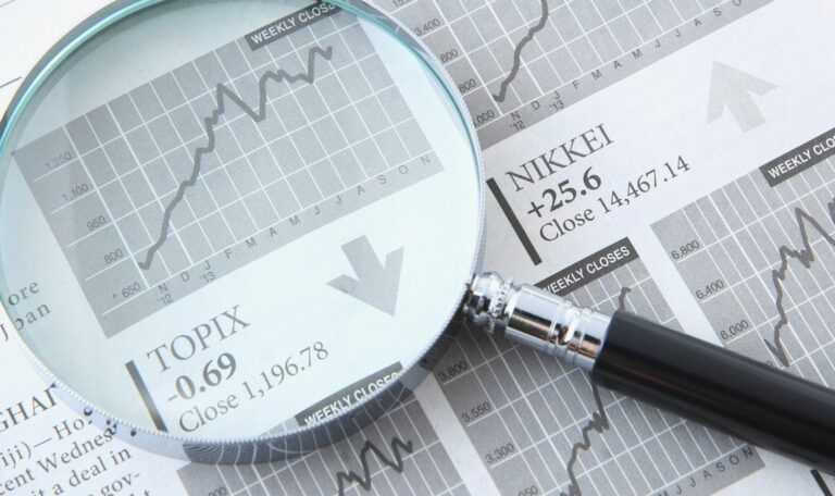 Gros plan sur une loupe grossissant un journal financier ouvert. Sous la loupe, un graphique boursier monte en flèche, avec un texte affichant l'indice 'TOPIX -0.69 Close 1,196.78' et une flèche pointant vers le bas, indiquant une tendance à la baisse locale. À côté, l'indice 'NIKKEI +25.6 Close 14,467.14' apparaît avec une flèche pointant vers le haut, symbolisant l'analyse des tendances économiques et des marchés financiers.
