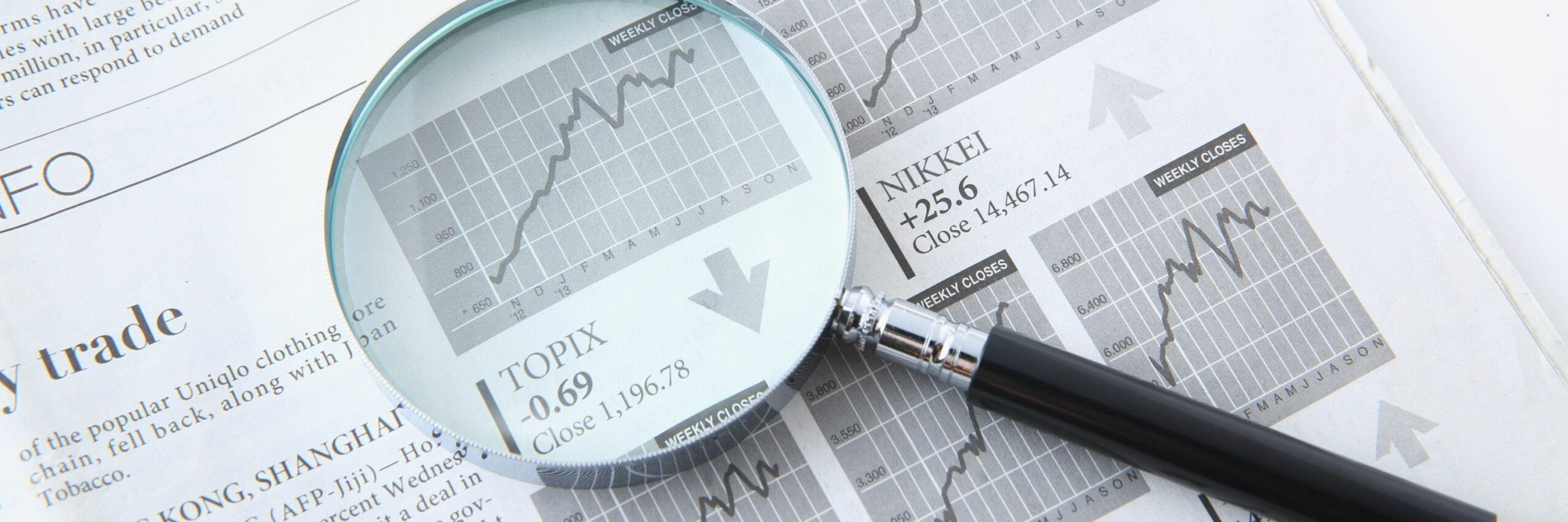 Gros plan sur une loupe grossissant un journal financier ouvert. Sous la loupe, un graphique boursier monte en flèche, avec un texte affichant l'indice 'TOPIX -0.69 Close 1,196.78' et une flèche pointant vers le bas, indiquant une tendance à la baisse locale. À côté, l'indice 'NIKKEI +25.6 Close 14,467.14' apparaît avec une flèche pointant vers le haut, symbolisant l'analyse des tendances économiques et des marchés financiers.